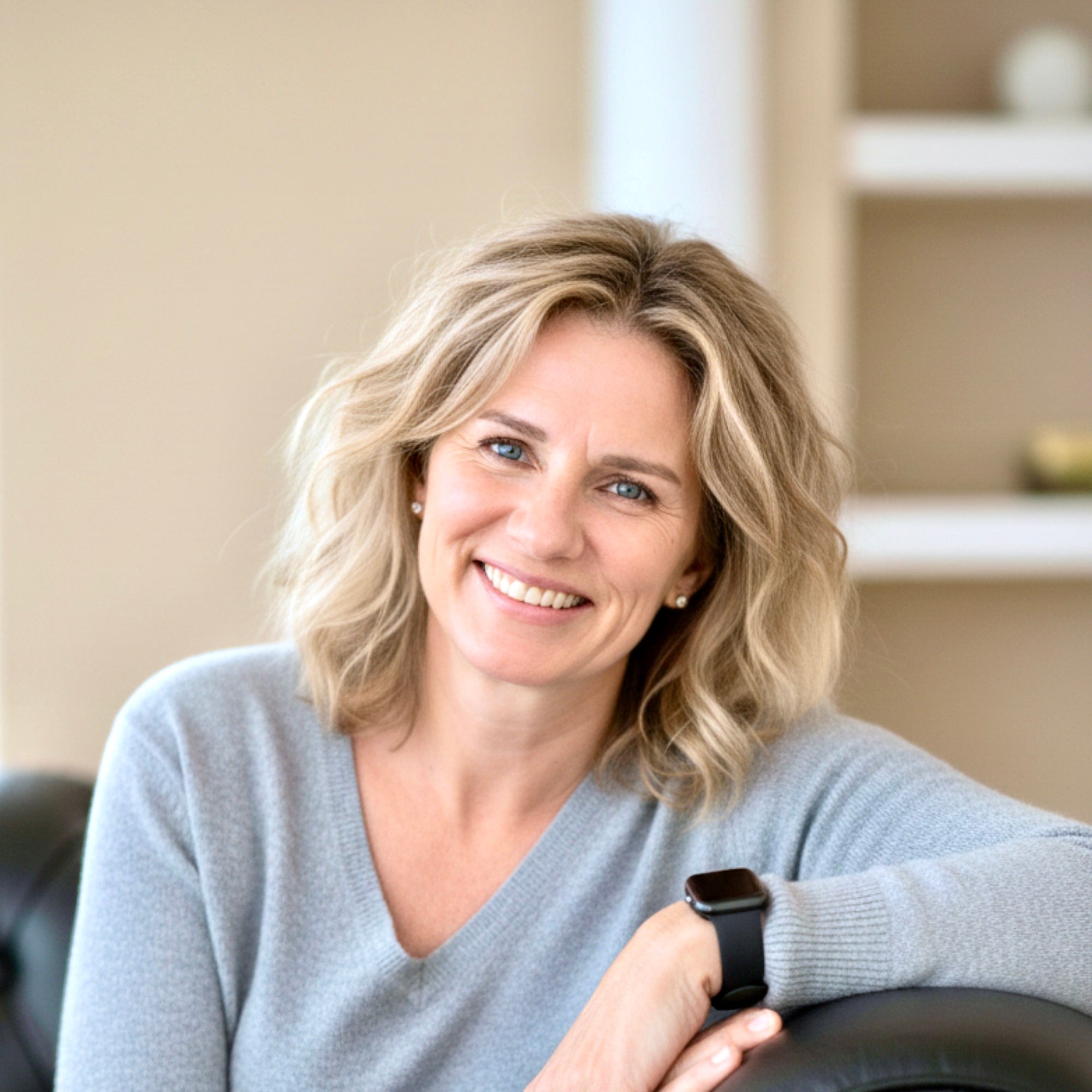Woman seated wearing a gray sweater holding a smartwatch indoors.