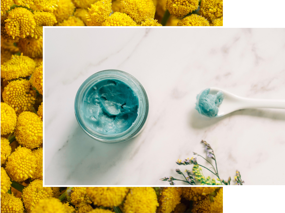 Jar of blue cream with a spoonful on a marble surface, surrounded by yellow flowers.