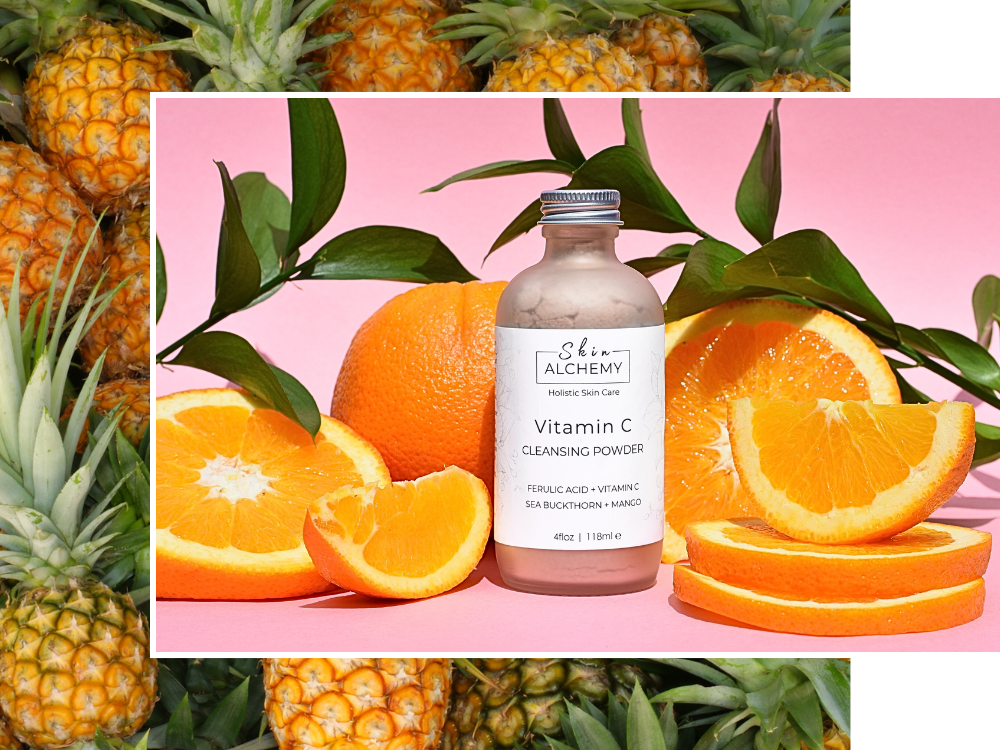 Bottle of 'Shea Alchemy' Vitamin C Cleansing Powder surrounded by oranges and pineapples on a pink background